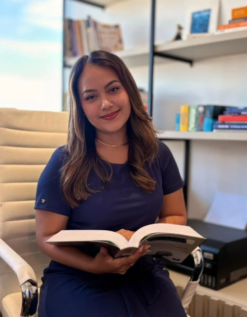 Giovana Tanaka sentada em seu consultório, com um livro aberto nas mãos e uma estante de livros ao fundo.
